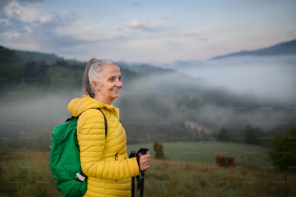 Uma mulher idosa caminhando na natureza no início da manhã com neblina e montanhas ao fundo.
