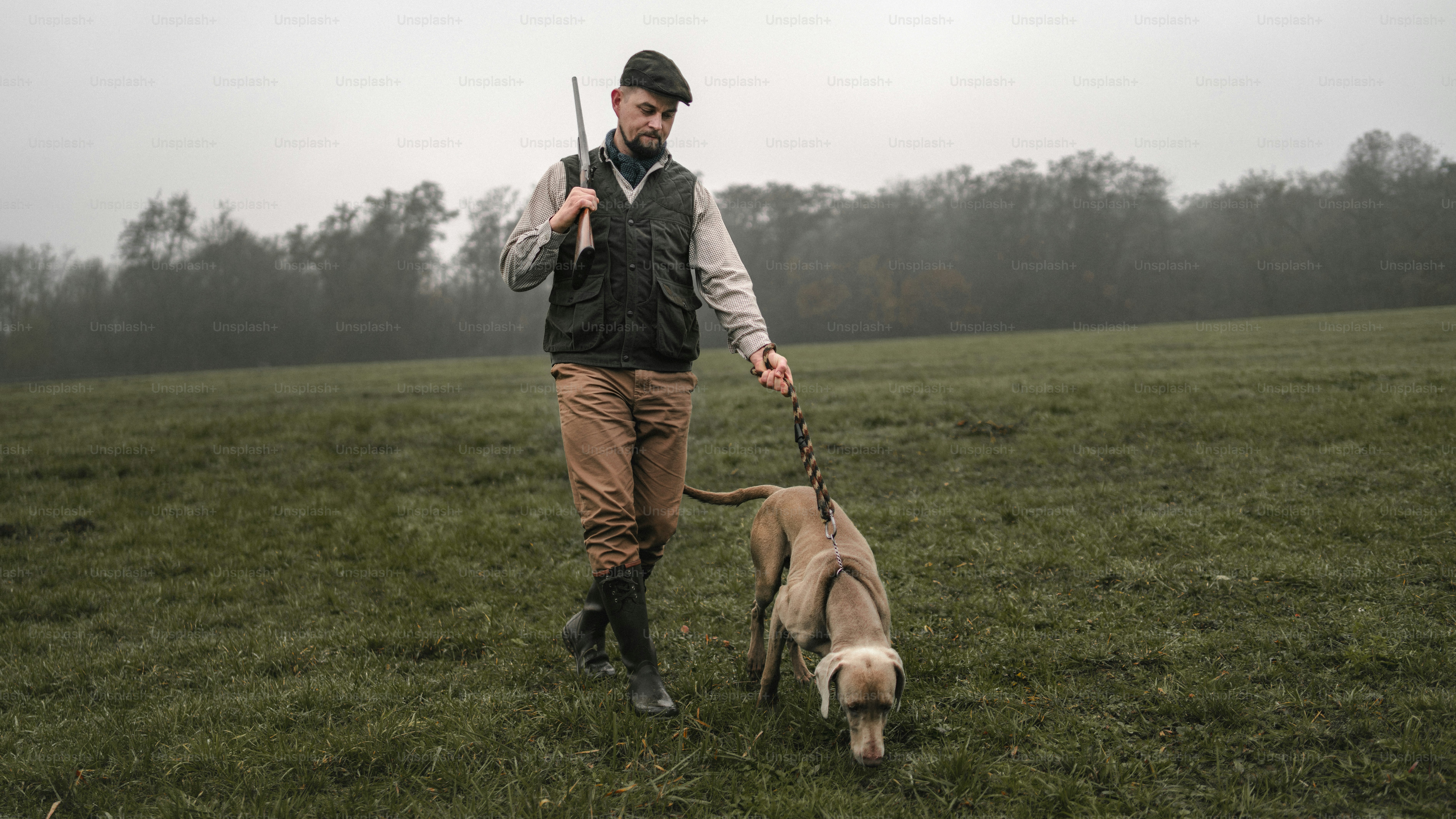 Un cazador con perro en ropa de tiro tradicional en el campo sosteniendo una escopeta.