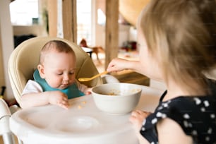 Two little children at home, unrecognizable toddler girl feeding her baby brother using spoon.