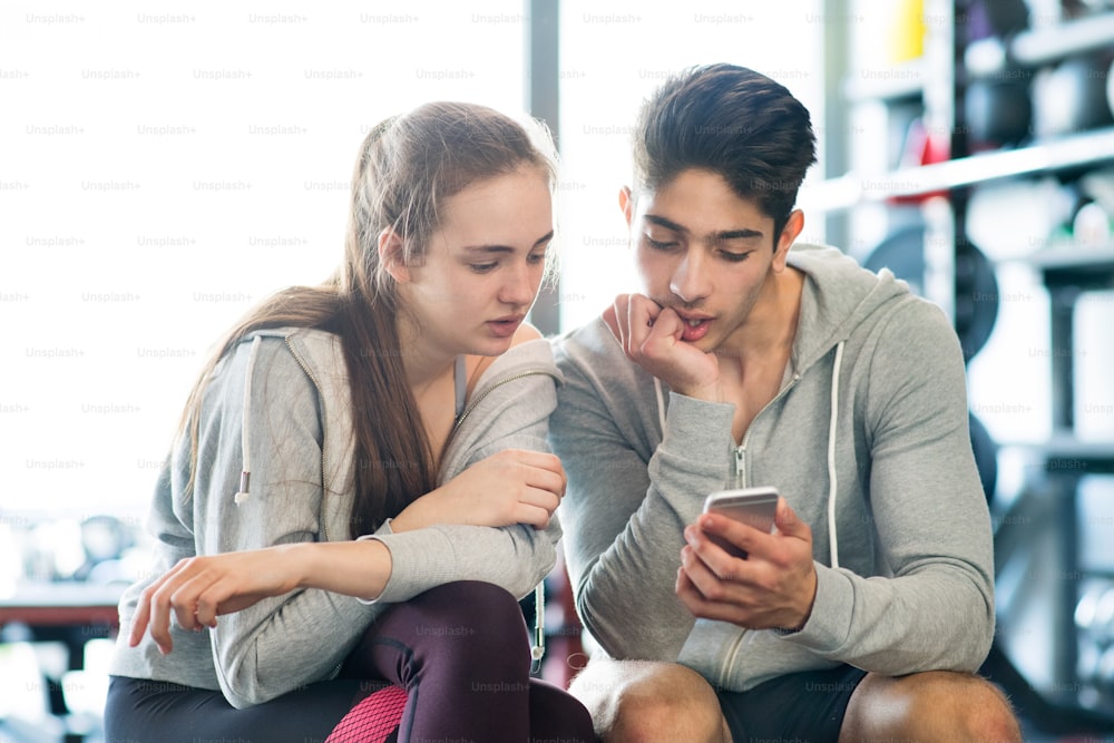 Beautiful young fit couple in modern gym gym talking, resting, holding smart phone, watching or writing something.