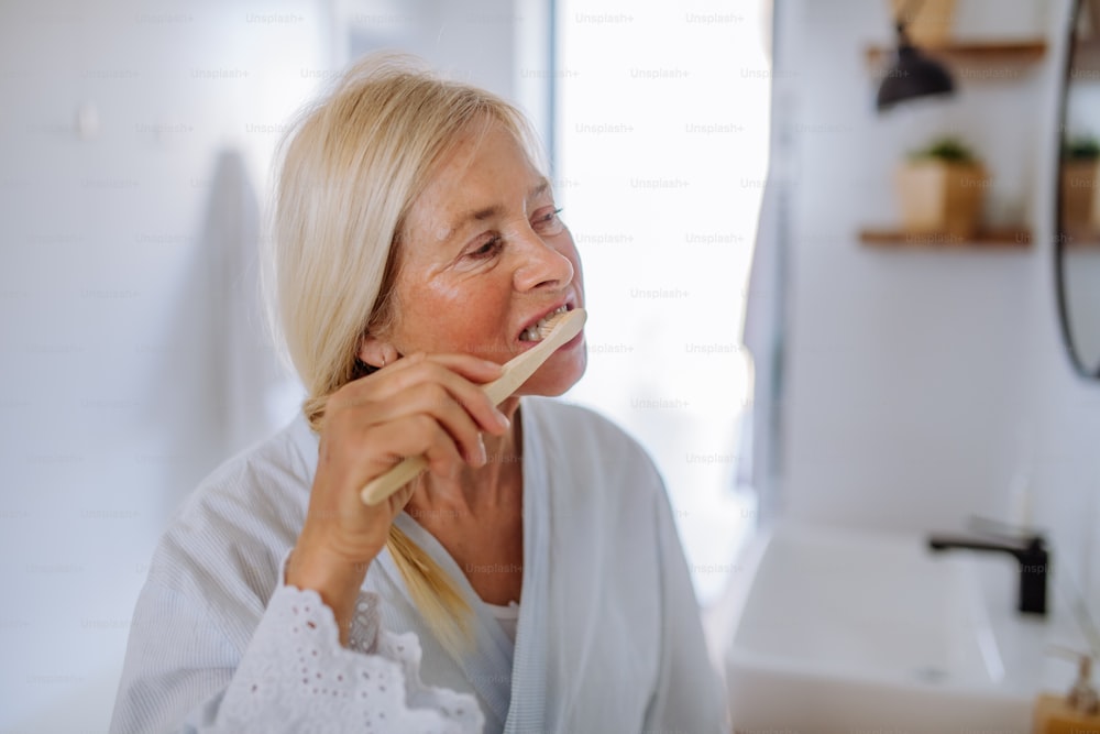 Une femme âgée en peignoir se brossant les dents avec une brosse à dents en bois écologique dans la salle de bain, un mode de vie durable.
