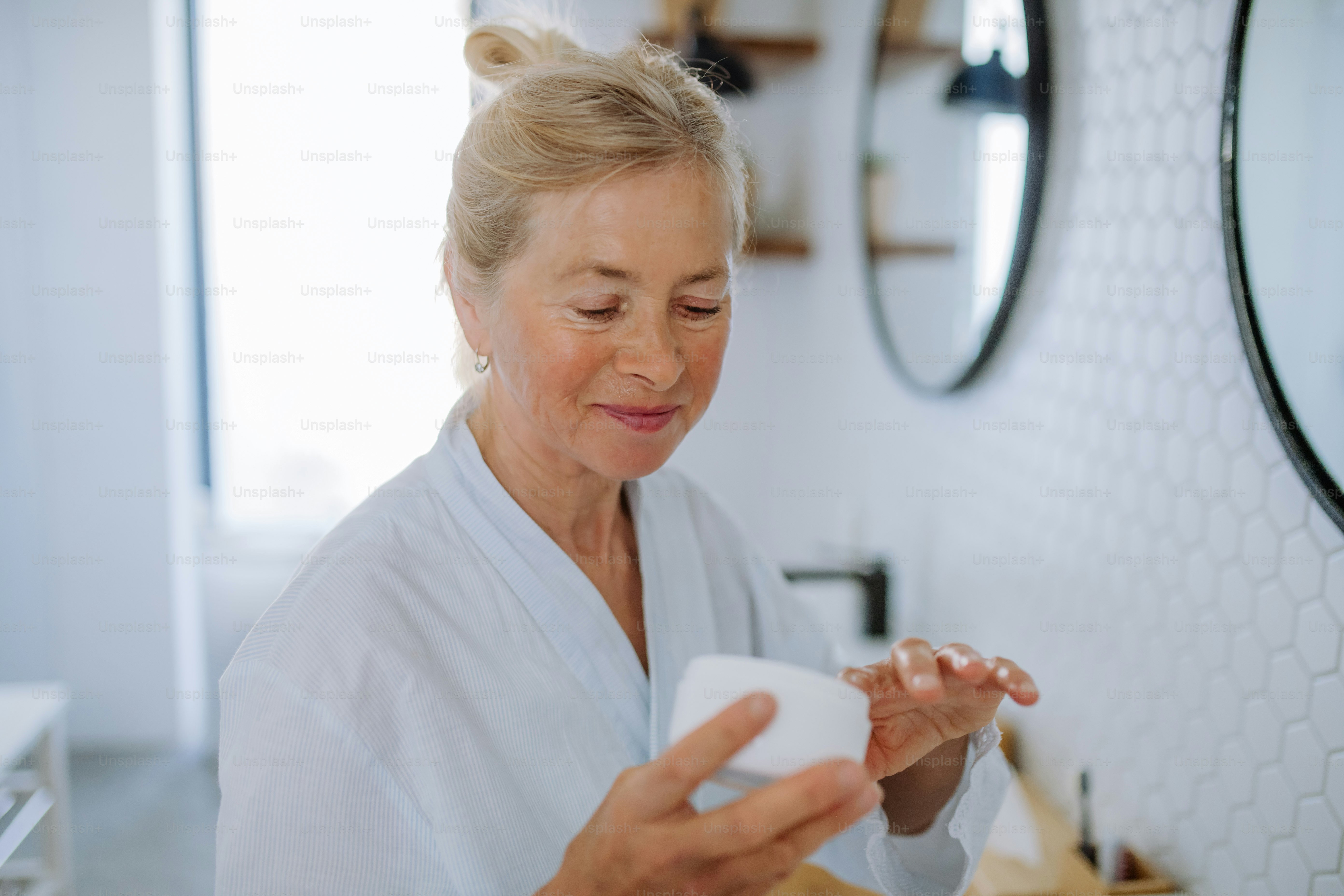 Uma bela mulher idosa em roupão aplicando creme natural no banheiro, cuidados com a pele e conceito de rotina matinal