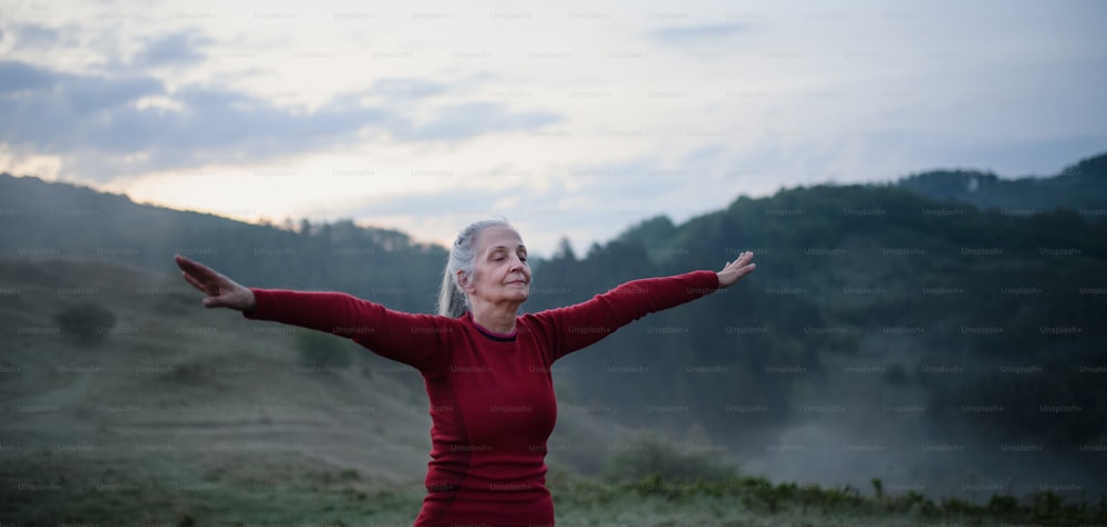 Une femme âgée faisant des exercices de respiration dans la nature tôt le matin avec du brouillard et des montagnes en arrière-plan.