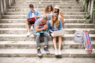 Group of attractive teenage students sitting on stone steps in front of university with laptop and notebooks, reading and studying.
