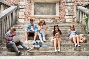 Group of attractive teenage students sitting on stone steps in front of university holding tablet, laptop and smart phones, reading or watching something.