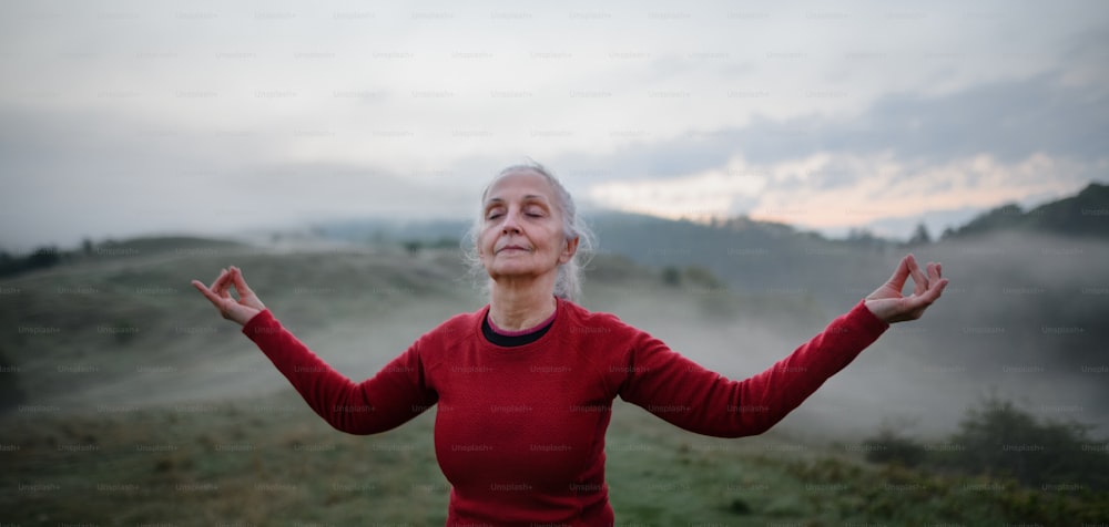 Una donna anziana che fa esercizio di respirazione nella natura la mattina presto con nebbia e montagne sullo sfondo.