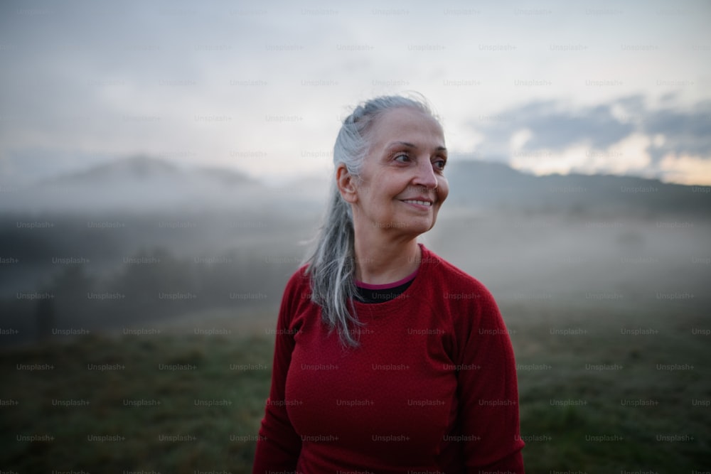 Eine fröhliche Seniorin in der Natur am frühen Morgen mit Nebel und Bergen im Hintergrund.