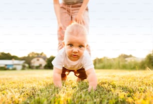 Cute baby boy with his unrecognizable mother crawling outside on the green grass.