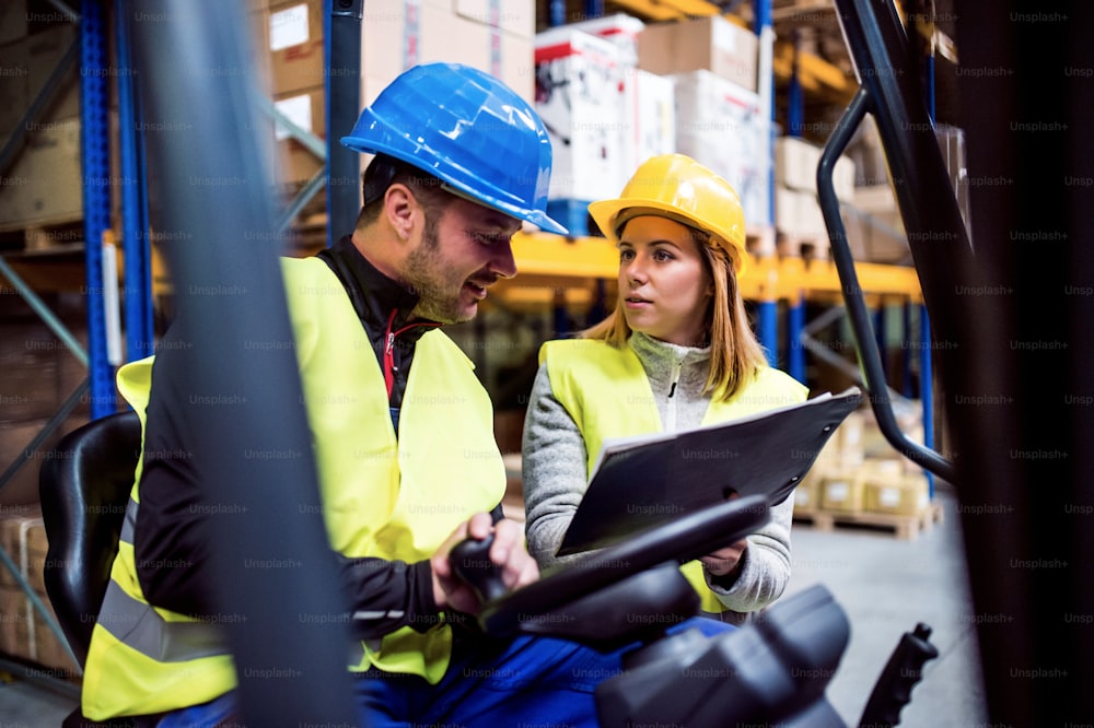 Young warehouse workers working together. Man sitting in a forklift and woman holding notes, discussing something.