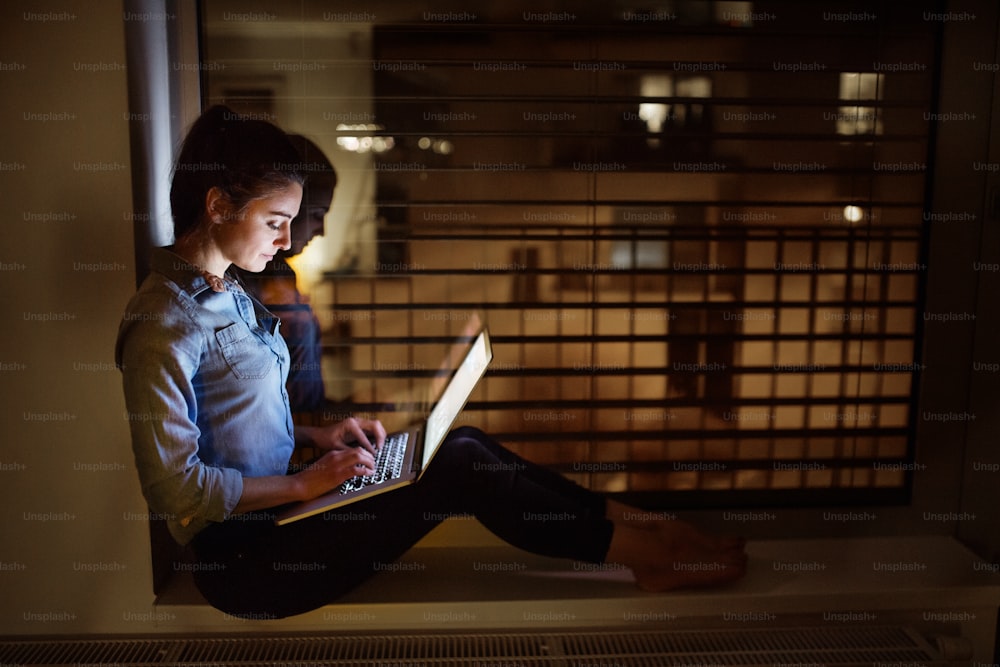 A woman sitting on a window sill at night, working on a laptop.