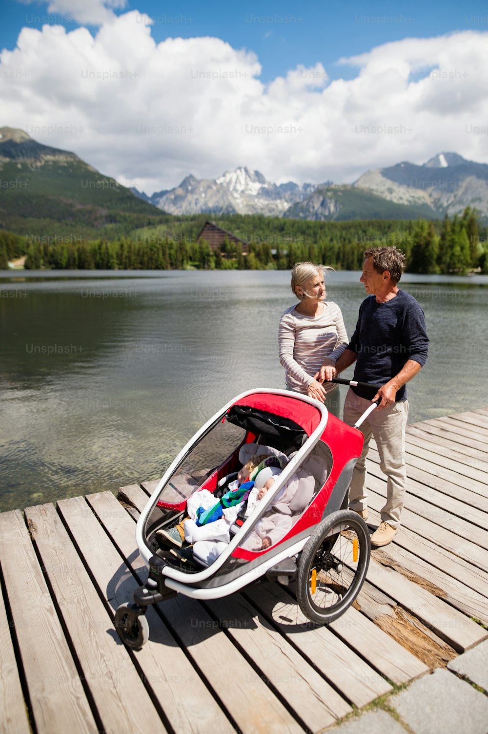 Seniorenpaar und Enkelkinder im Jogging-Kinderwagen, Sommertag. Hohe Berge im Hintergrund.