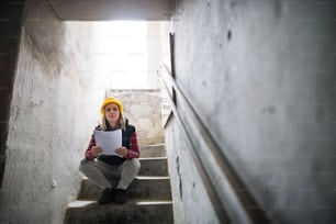 Female worker on the building site. Beautiful young woman sitting on stairs, holding blueprints. House construction