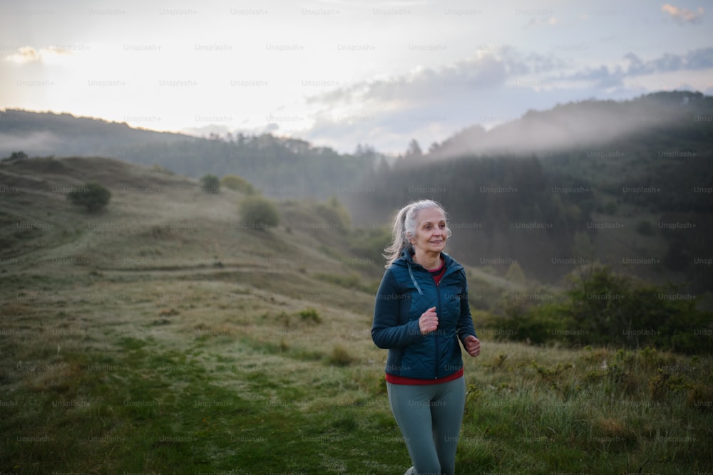 A senior woman jogging in nature on early morning with fog and mountains in background.