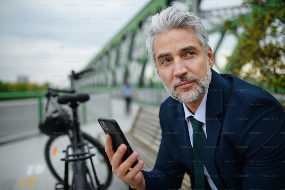 A businessman with bike sitting on bench, using smartphone. Commuting and alternative transport concept