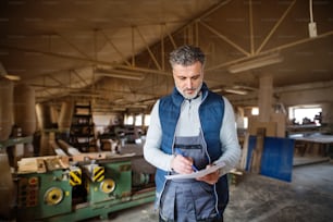 A mature man worker in the carpentry workshop, holding paper and a pen, making plans.