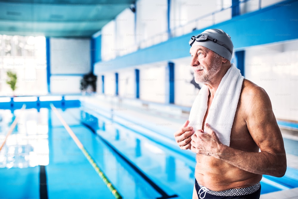 Senior man standing by the indoor swimming pool, arms on hips. Active pensioner enjoying sport.