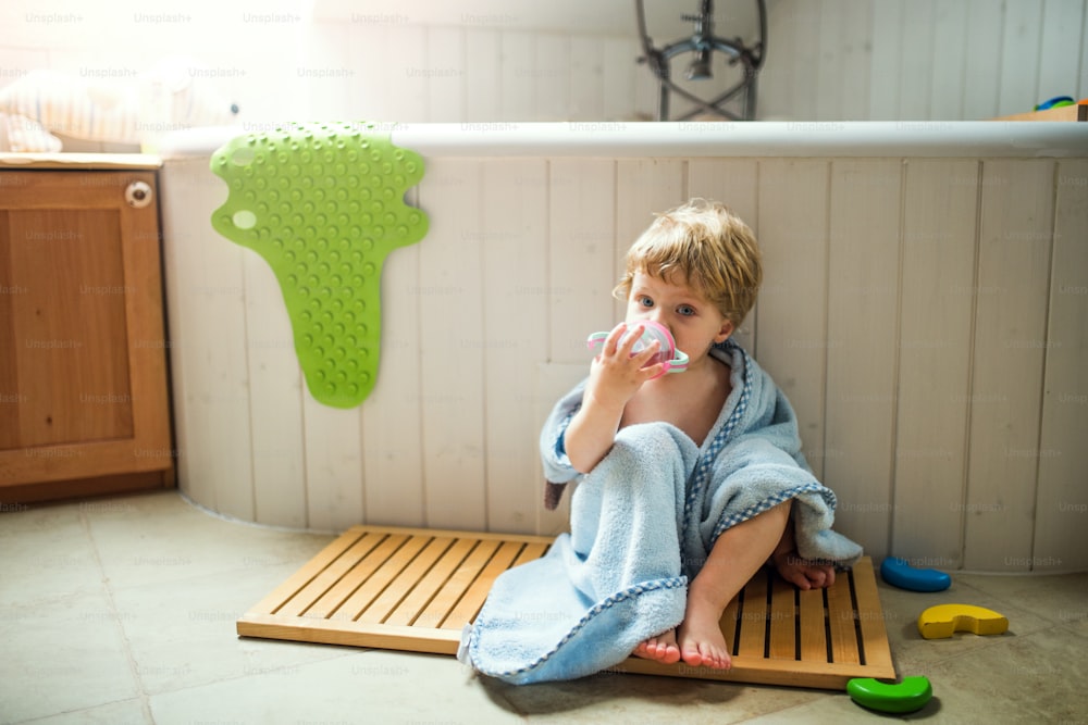 A toddler boy wrapped in towel sitting on the floor in the bathroom at home, drinking.