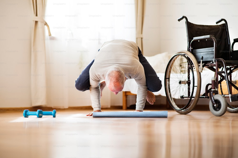 Active senior man doing exercise at home. Man next to a wheelchair doing frogstand. Knees on elbows.