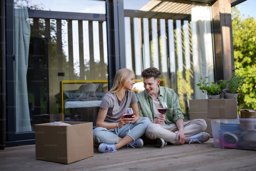 Um jovem casal alegre que se muda em sua nova pequena casa em bosques sentados no chão do terraço e bebendo vinho. Concepção de vida móvel e sustentável.