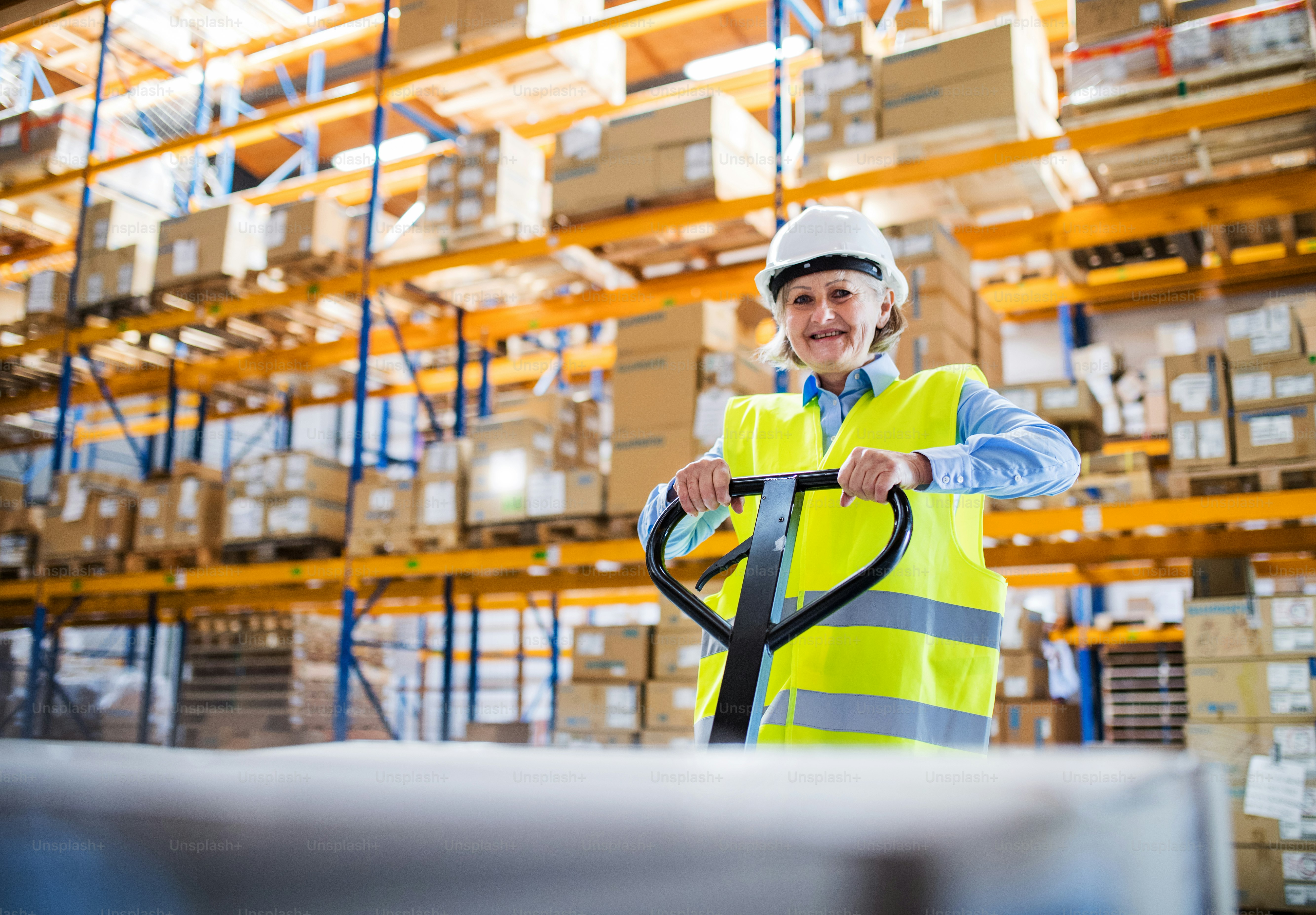 A senior woman warehouse worker or supervisor pulling a pallet truck with boxes.