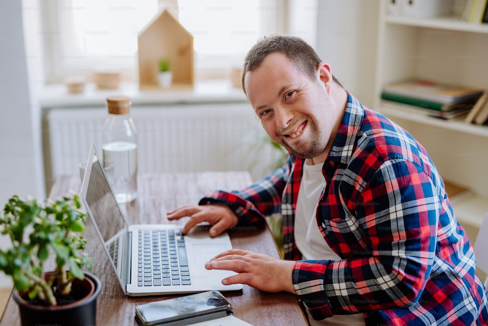 A young man with Down syndrome sitting at desk at home and using laptop, looking at camera and smiling.