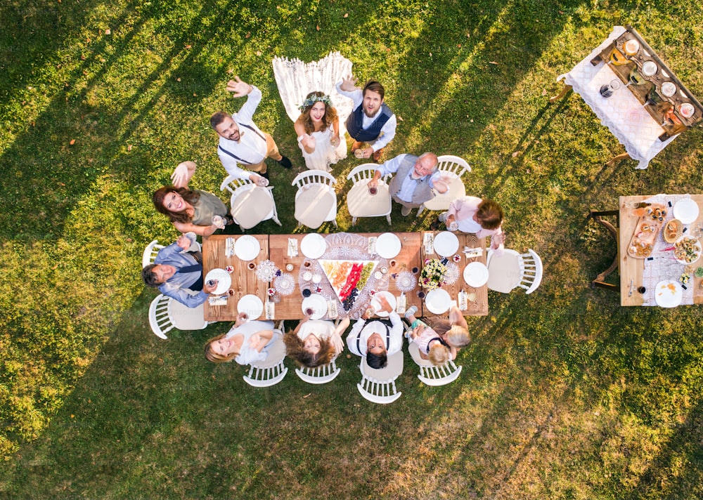 Wedding reception outside in the backyard. Bride and groom with a family standing around the table, waving. Aerial view.