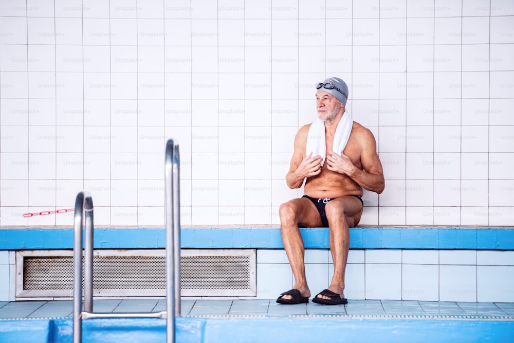 Senior man sitting by the indoor swimming pool, towel around his neck. Active pensioner enjoying sport.
