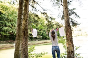 Beautiful woman on camping holiday in forest at the lake hanging clothes, rear view.