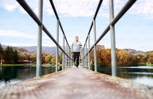 Young athlete with smartphone in gray jacket running outside. Trail runner training for cross country running in colorful sunny autumn nature.