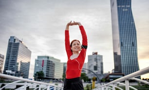 Front view of young woman runner with earphones in city, stretching on the bridge.
