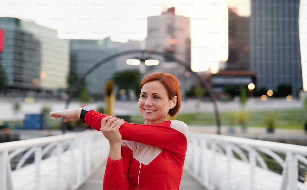 Front view of young woman runner with earphones in city, stretching on the bridge.