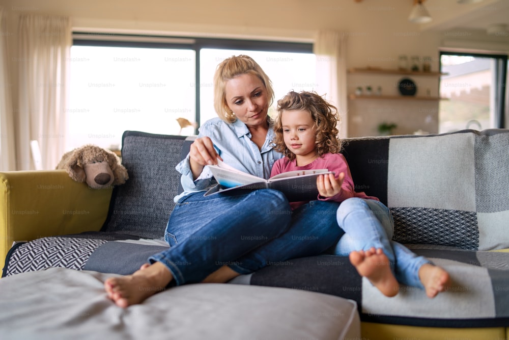 Une jolie petite fille avec sa mère assise sur un canapé à l’intérieur à la maison, lisant un livre d’histoires.