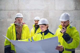 建設現場のコンクリートの壁に向かって立ち、設計図を手にするエンジニアのグループ。