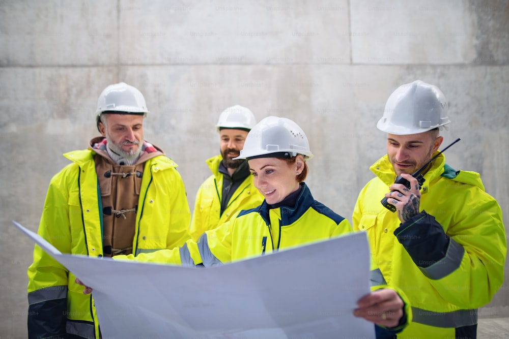 Un grupo de ingenieros de pie contra un muro de hormigón en una obra de construcción, sosteniendo planos.