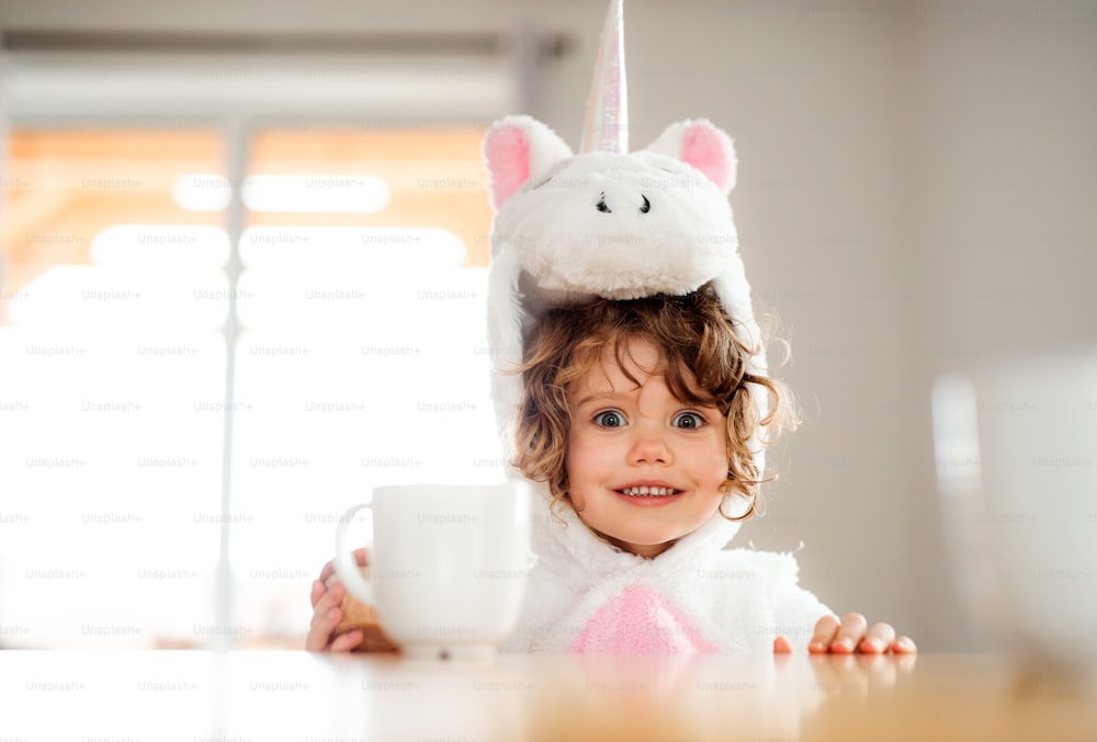 A portrait of happy small girl in unicorn mask sitting at the table at home.