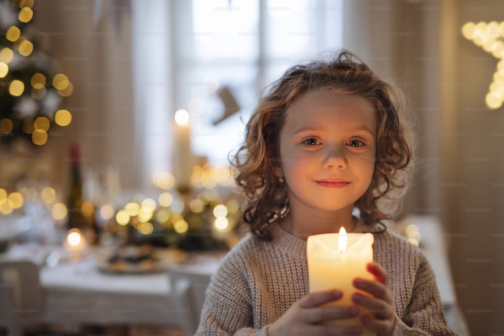 Front view of cheerful small girl standing indoors at Christmas, holding candle.