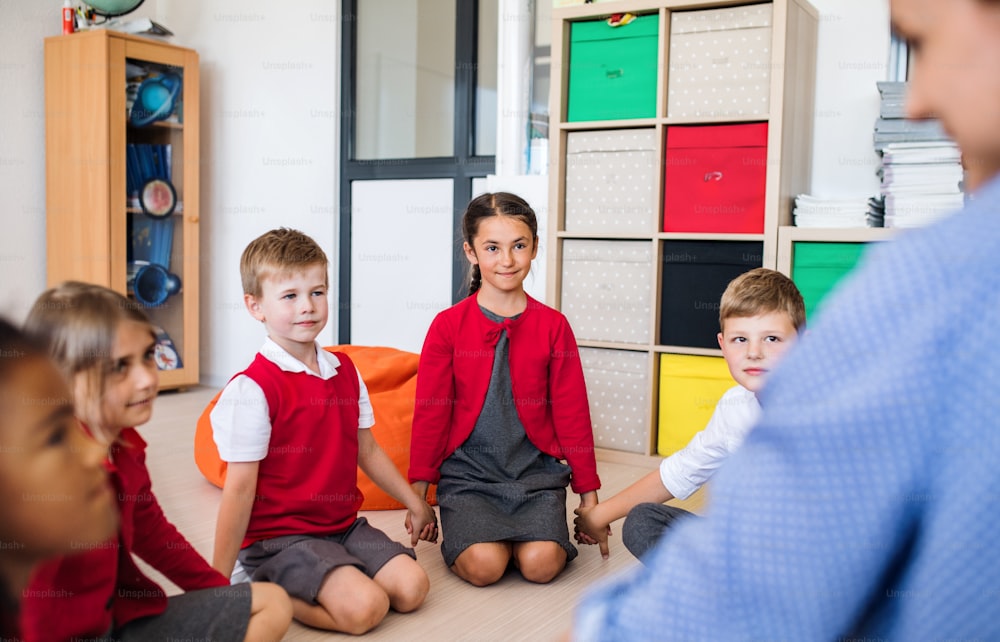 Um grupo de pequenos alunos com um professor irreconhecível sentado no chão em sala de aula, de mãos dadas.