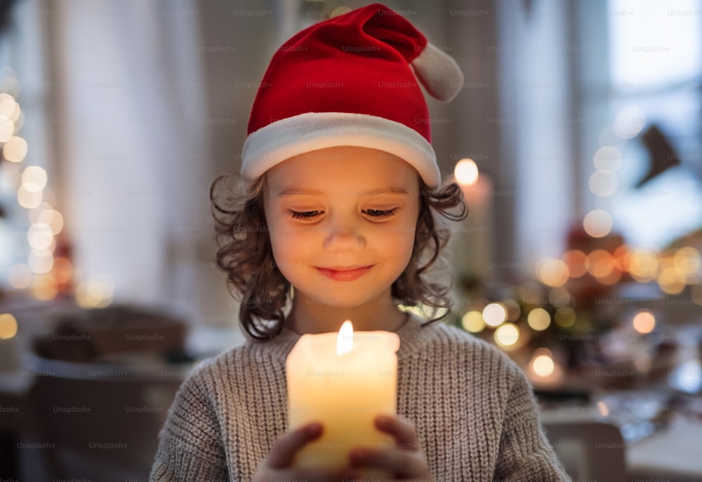 Vista frontal de una niña pequeña alegre con sombrero de Papá Noel en el interior en Navidad, sosteniendo una vela.