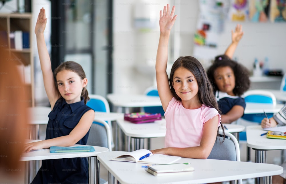 Pequenas crianças em idade escolar sentadas na mesa da sala de aula na lição, levantando as mãos.