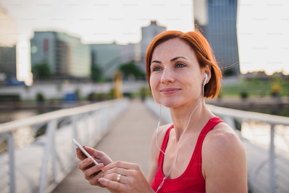 Corredora de mulher nova com fones de ouvido na cidade, usando smartphone ao descansar.