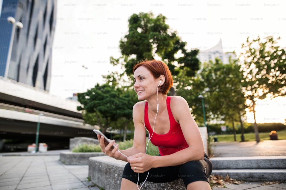 Young woman runner with earphones in city, using smartphone when resting.