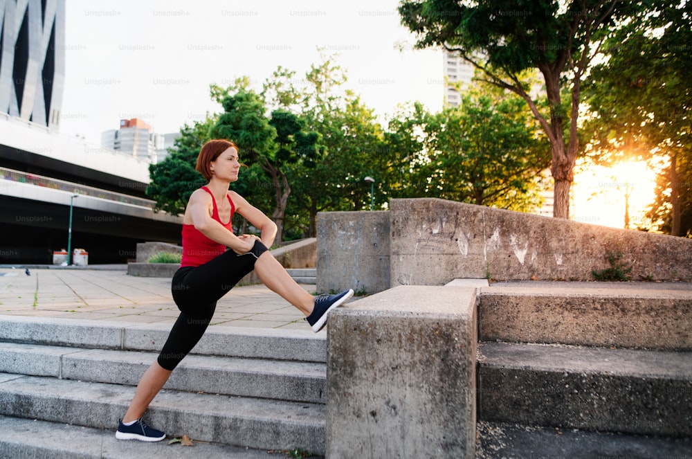 A young woman doing exercise outdoors in city at sunset, stretching.