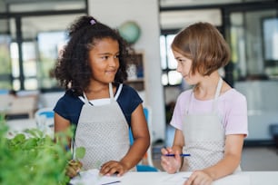 Two small school kids with aprons standing at the desk in class, talking.