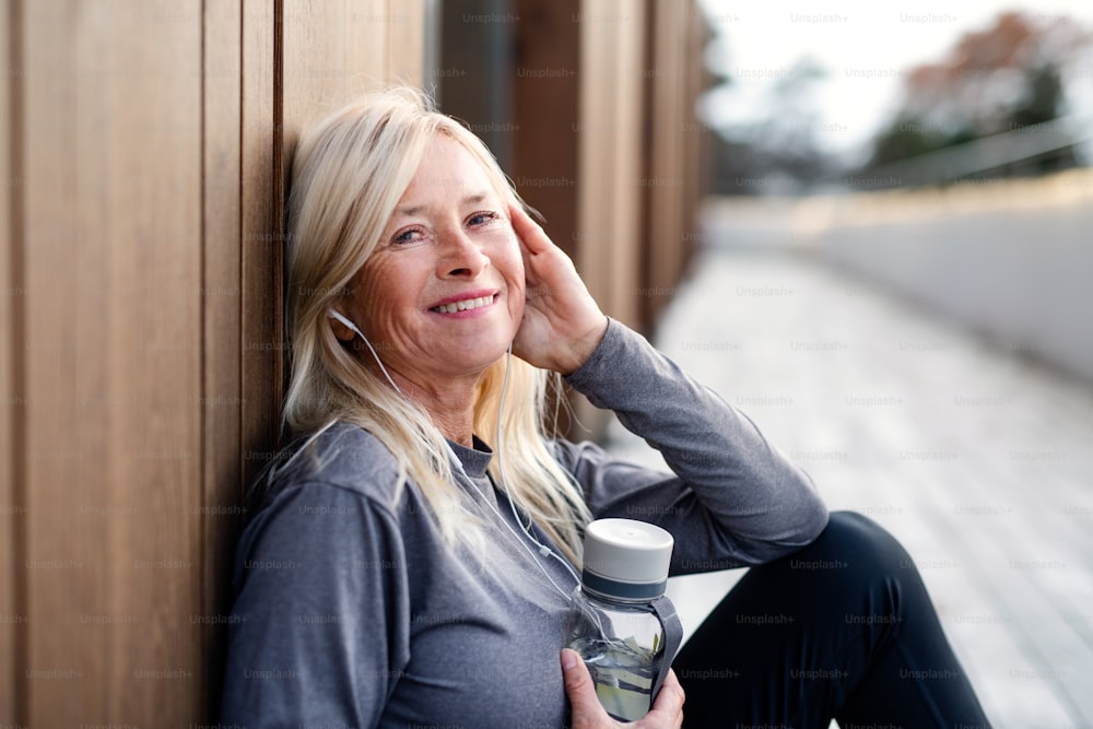 A side view of senior woman with earphones sitting outdoors resting after exercise.