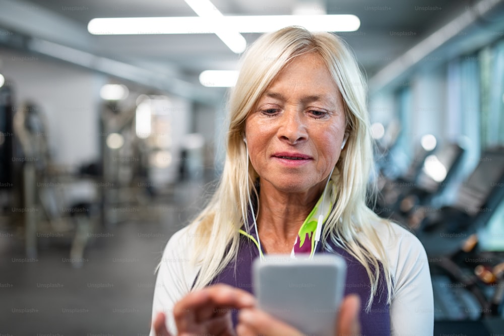 A front view of senior woman with earphones and smartphone in gym resting after doing exercise.