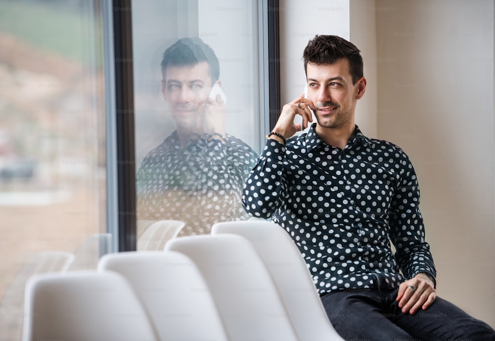Un joven con un teléfono inteligente sentado junto a la ventana, haciendo una llamada telefónica. Espacio de copia.