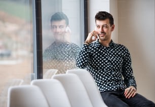 A young man with smartphone sitting by the window, making phone call. Copy space.