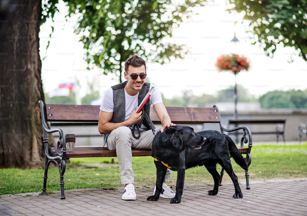Junger blinder Mann mit weißem Stock und Blindenhund sitzt auf der Bank im Park in der Stadt und ruht sich aus.