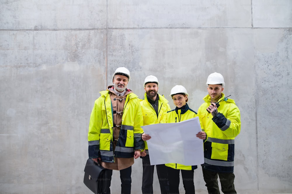 Un grupo de ingenieros de pie contra un muro de hormigón en una obra de construcción, sosteniendo planos. Espacio de copia.