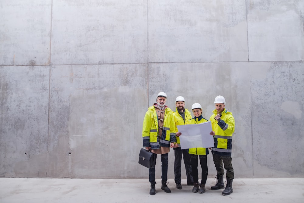Eine Gruppe von Ingenieuren steht an einer Betonwand auf der Baustelle und hält Baupläne in der Hand. Speicherplatz kopieren.